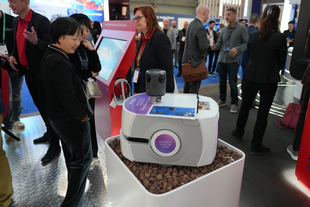 A group of people at a technology expo observe a white, boxy robot displayed on brown rocks in a white container. One woman gestures, others watch attentively, and interactive screens and exhibition booths are visible in the background.