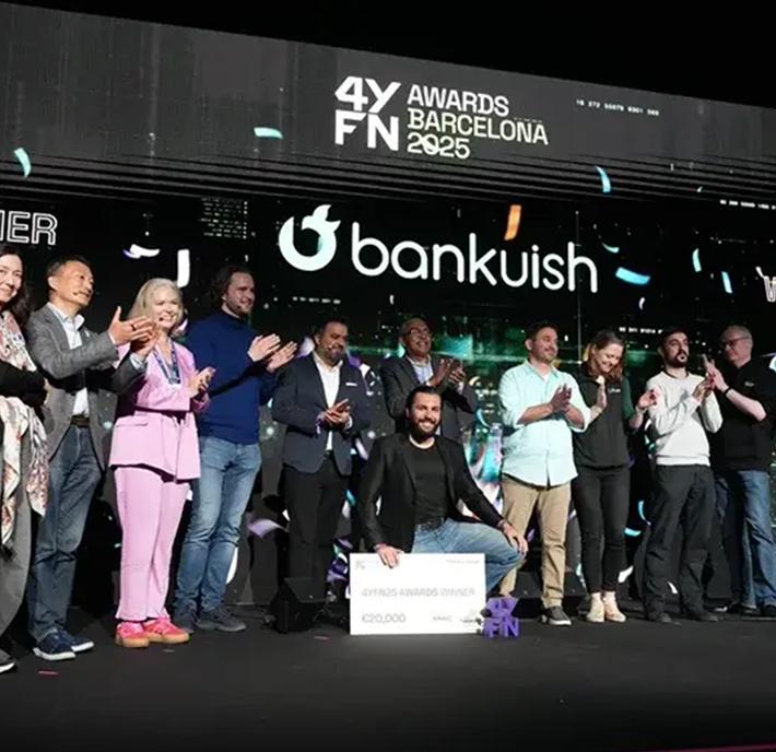 A group of people stand on stage, smiling and applauding as confetti falls, with one person kneeling in front holding a large check. The backdrop reads “4YFN Awards Barcelona 2025” and “bankuish,” celebrating a triumphant 4YFN award presentation.