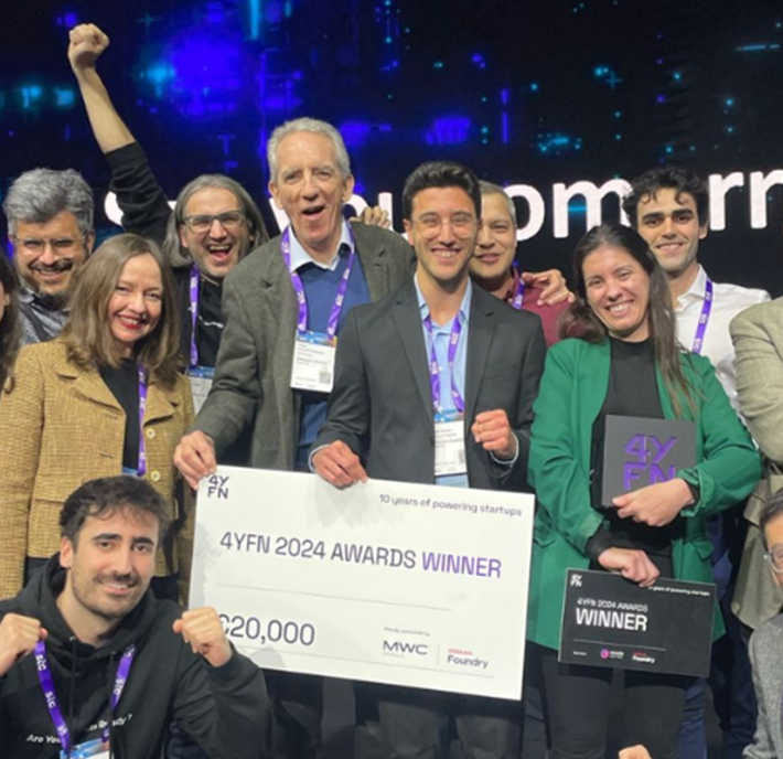 A group of joyful people pose onstage at the 4YFN event, holding a large winner’s check for €20,000 labeled “4YFN 2024 Awards Winner.” Several wear lanyards and badges; one holds a trophy before a lit display with event branding.