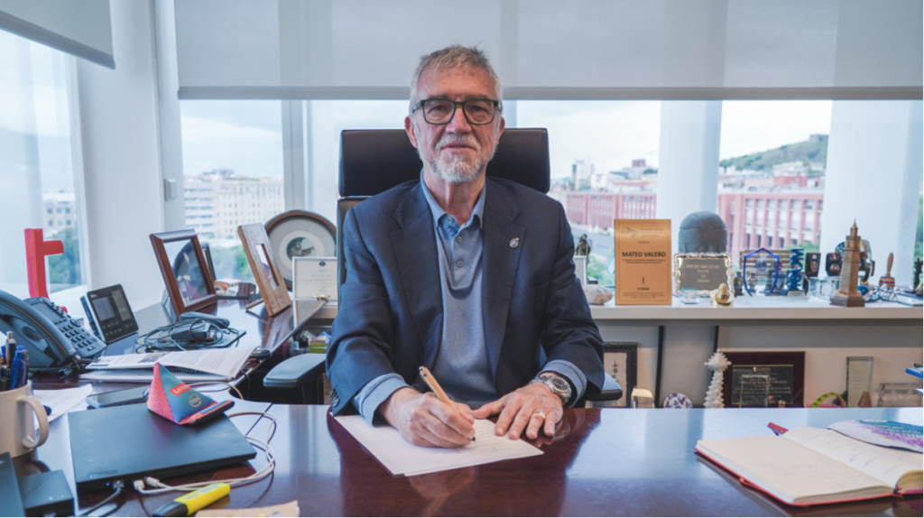An older man with grey hair and glasses sits at a large desk in an office, holding a pen over a sheet of paper. He wears a dark blazer and blue shirt. The desk is filled with books, awards, and materials from the Barcelona Supercomputing Centre.