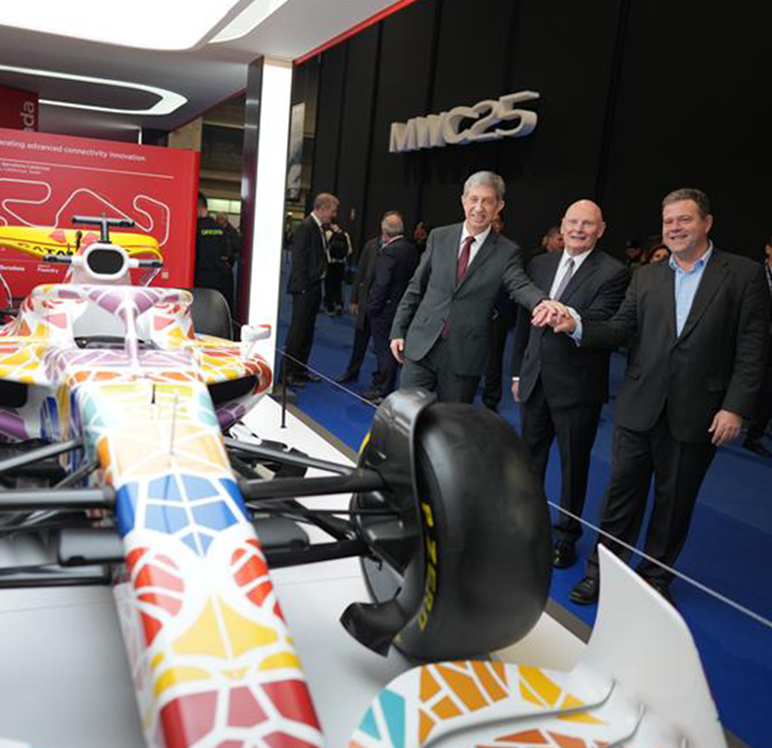 Three men in suits stand smiling and shaking hands next to a white Formula 1 racing car with a colourful geometric pattern. The event, held indoors, features a large red CircuitX display showcasing a racing track diagram in the background.