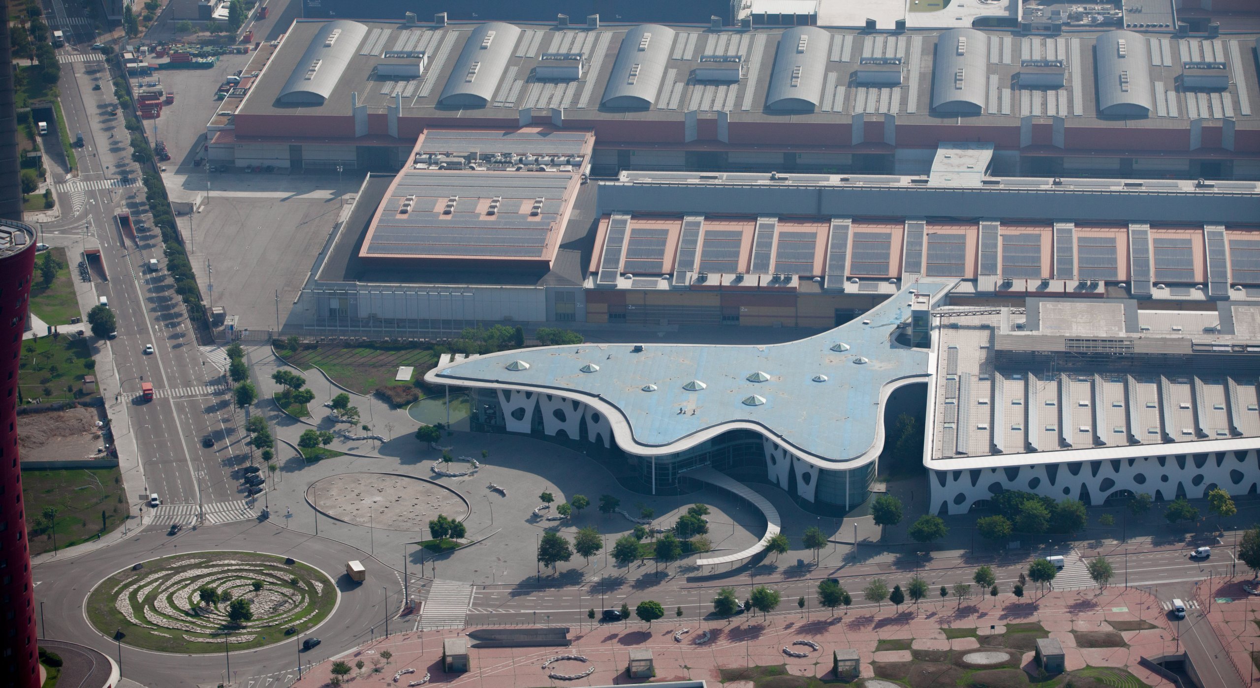 Aerial view of a modern building with a blue, star-shaped roof and white walls featuring irregular black oval windows. Surrounding roads, a roundabout with a small garden, and adjacent rectangular industrial buildings are visible. Trees line some streets.