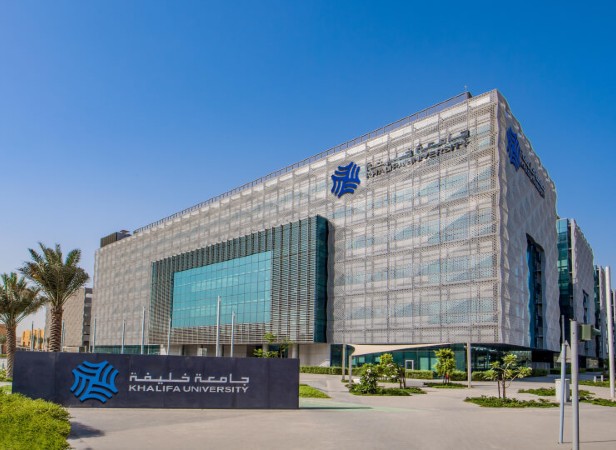 Modern Khalifa University building with a sleek, glass facade, patterned metal exterior, and blue logos in Arabic and English. Palm trees and green landscaping surround the structure under a clear blue sky. A bold sign with the university’s name stands in front.