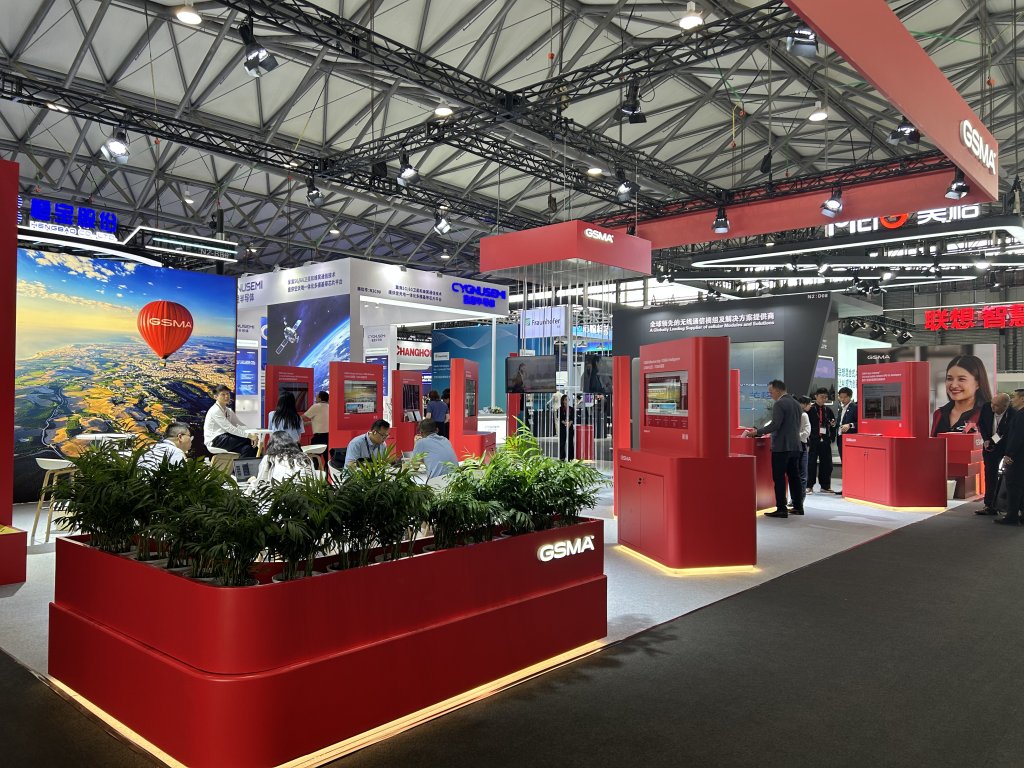 Exhibition hall with red GSMA booths, digital displays, plants, and people interacting under bright lights.