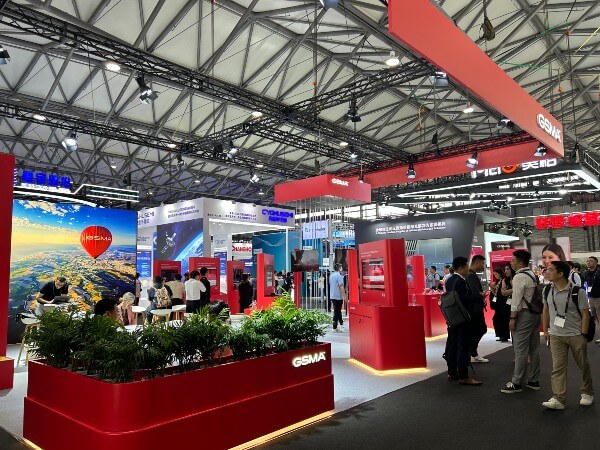 A bustling tech expo hall features bright red GSMA booths, digital displays, greenery, and groups of people talking and exploring exhibits. Overhead lighting and signage add a modern feel to the vibrant, crowded scene.