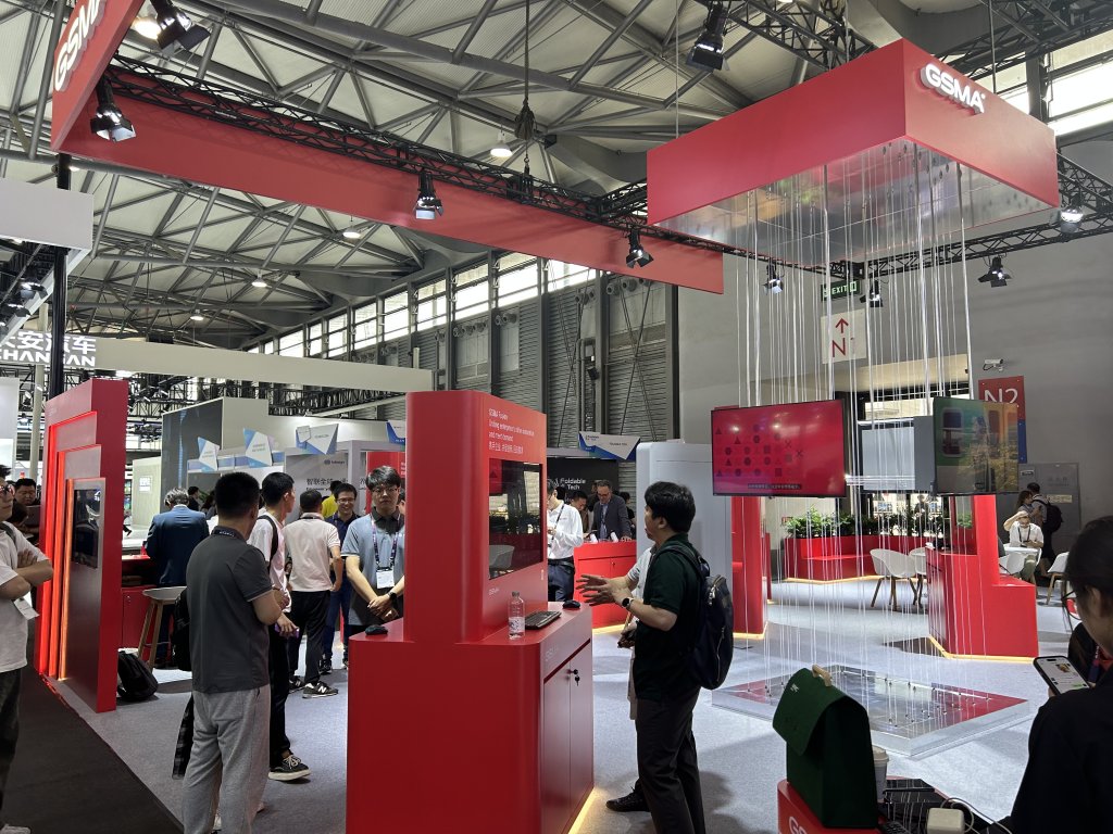 People explore a tech exhibition booth with red displays, screens, and a modern open setup under bright lighting.