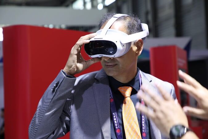 A man in a grey suit, orange tie, and conference lanyard tries on a white virtual reality headset indoors. He lifts one hand to the headset while another person's blurred hand gestures in the foreground. Red panels and bright lighting are in the background.