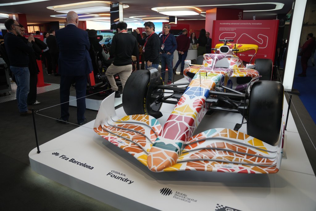 A colorful, geometric-patterned Formula 1 car is displayed indoors at an exhibition. People in business attire and badges gather around the car, engaging in conversation. Exhibition booths and banners are visible in the background.