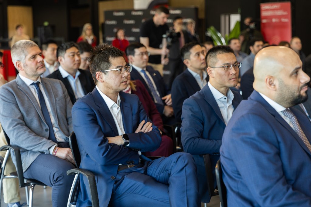A group of men in suits sit attentively in rows at a business conference or seminar.