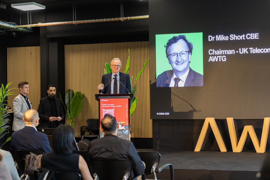 A man speaks at a podium in front of an audience with Dr. Mike Short's photo and title displayed on a screen.
