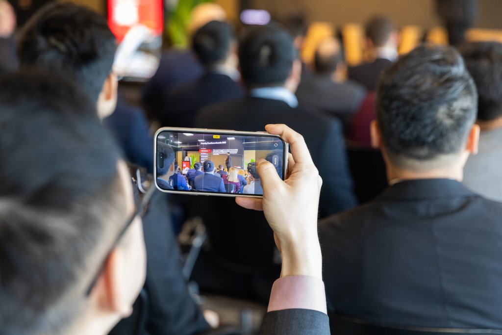 Person filming a conference or presentation on their phone among a seated audience.