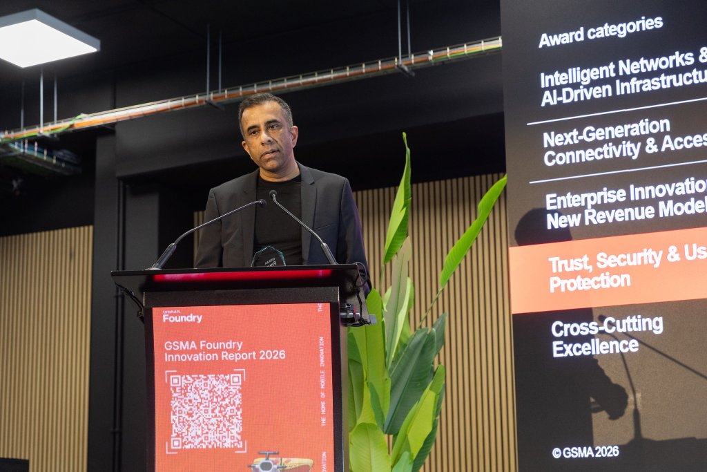 A man stands at a podium speaking at an event, with a screen listing award categories beside him.