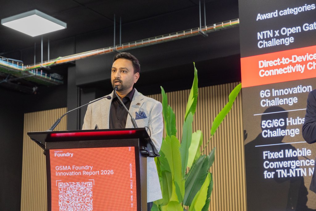 A man speaks at a podium during a tech event, with a digital screen listing award categories beside him.