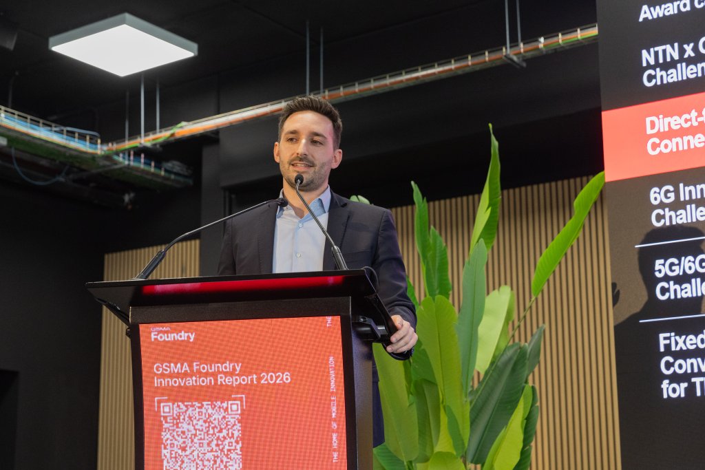 A man in a suit speaks at a podium displaying "GSMA Foundry Innovation Report 2026" in a modern venue.