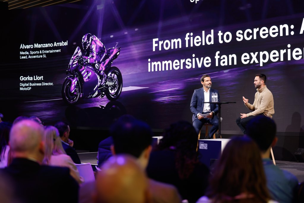 Two men speak on stage at a conference with a large screen showing MotoGP graphics behind them.