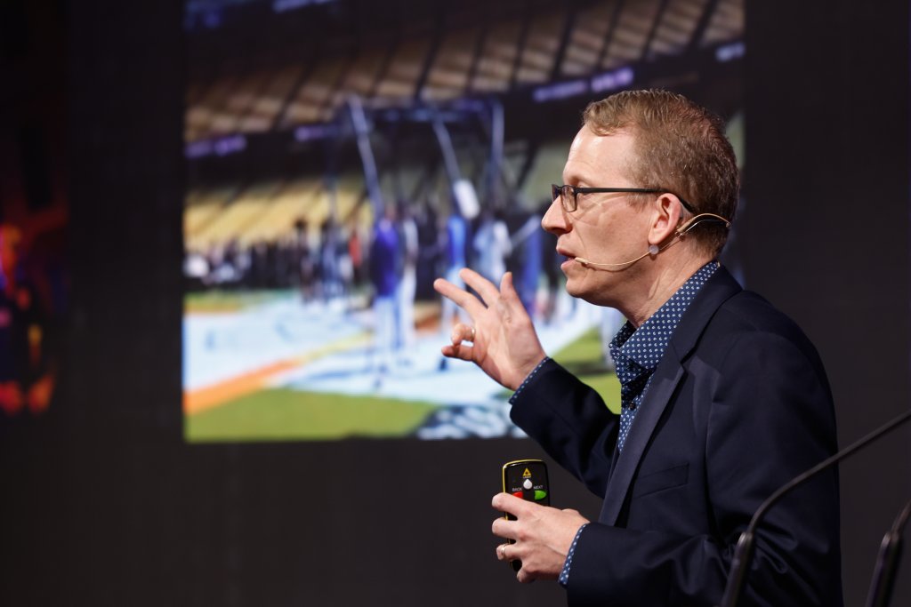 A man in a suit gives a presentation with a microphone, gesturing in front of a sports scene on a large screen.