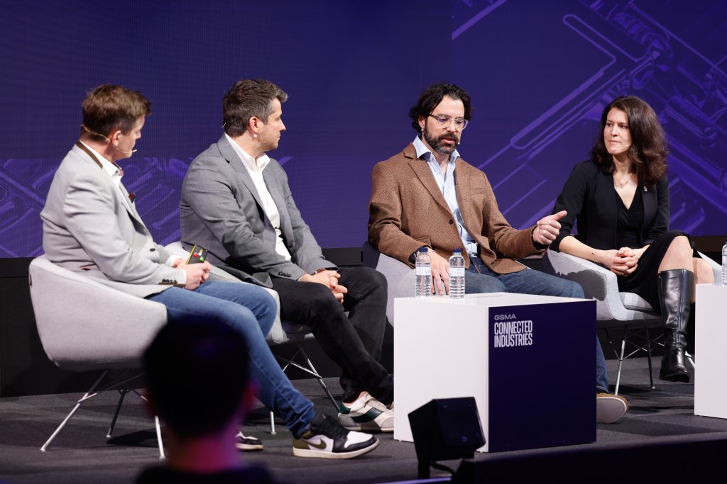 Four people sit on stage in discussion at a tech conference, with microphones and water bottles on tables.