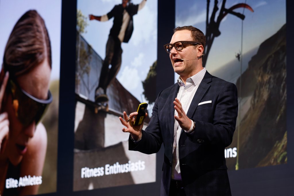 Man in suit giving a presentation, standing in front of large sports-themed images and text on a screen.