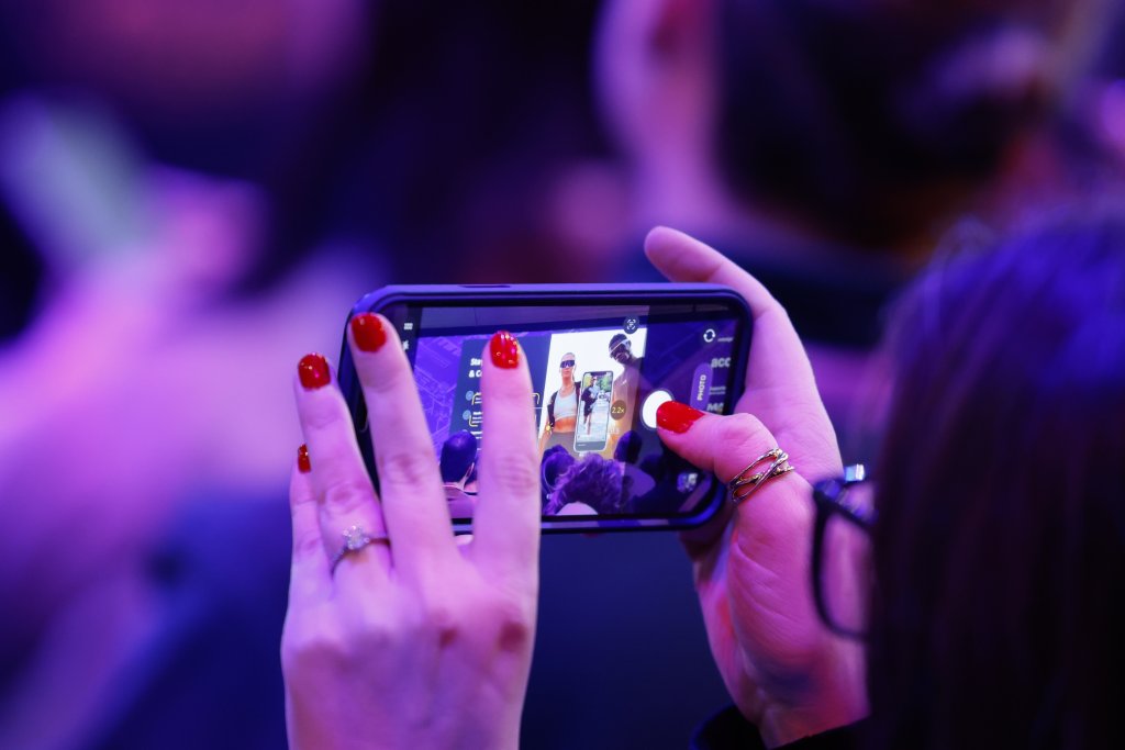 A person with red nails takes a photo of a performer on stage using their phone in a crowded setting.