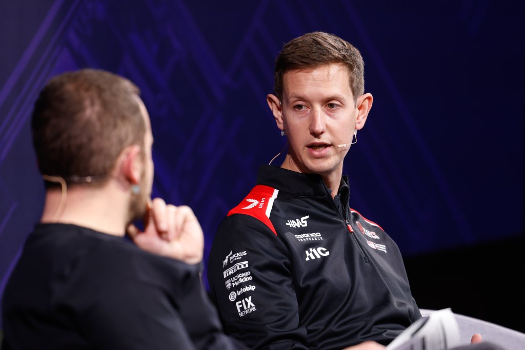 Two men with microphones have a discussion on stage; one wears a black sports jacket with logos.