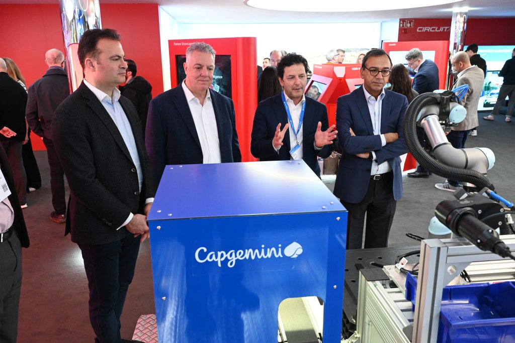 Four men observe a blue Capgemini machine at a tech event, with people and displays in the background.