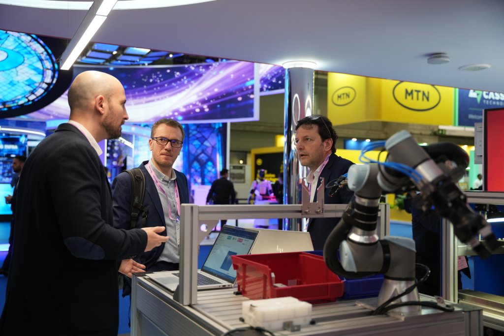 Three men talk at a tech event, standing near a robotic arm with screens and bright lights in the background.