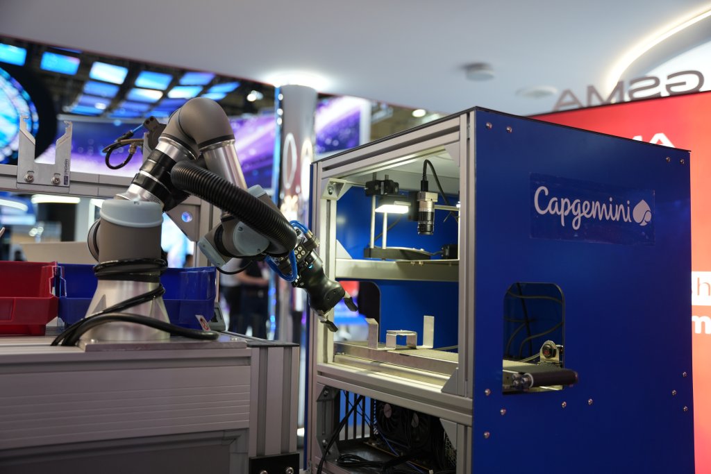 A robotic arm works inside a blue Capgemini-branded enclosure at a technology exhibition.