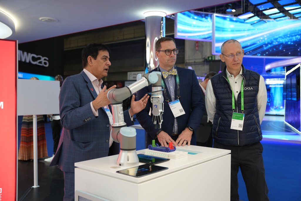 Three men observe a robotic arm demonstration at a technology exhibition, with colorful digital displays behind them.