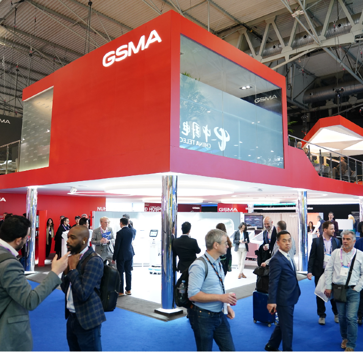 People networking and walking at a busy GSMA event booth with a red and white modern design, featured in the Q1 2026 GSMA Foundry Newsletter.