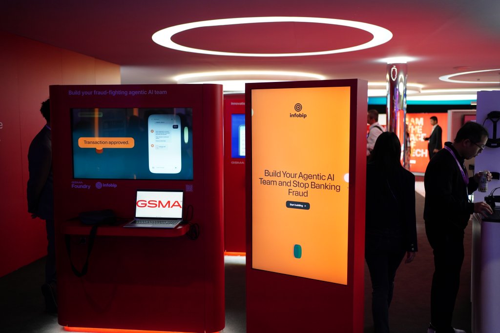 People stand near bright orange AI fraud prevention displays at a tech event, with screens and signage visible.