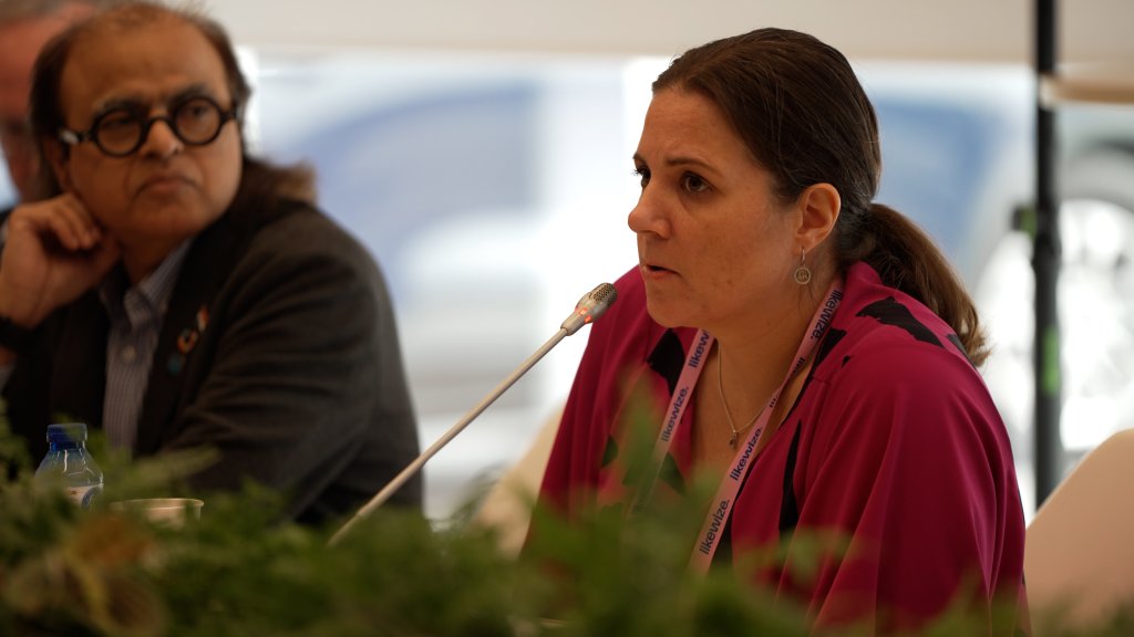 A woman speaks into a microphone at a conference, with a man seated beside her and plants in the foreground.