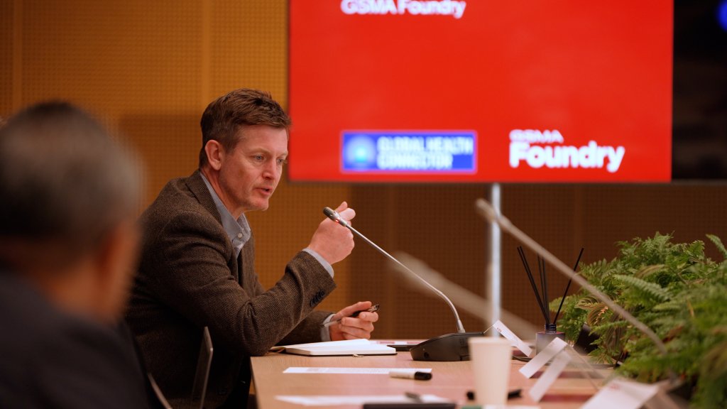 A man speaks into a microphone at a conference table with a red GSMA Foundry screen in the background.