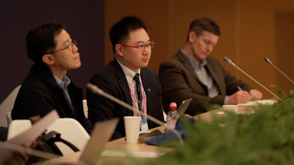Three men in suits sit at a conference table, listening attentively and taking notes.