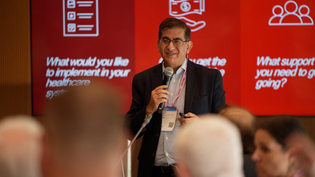 A man in a suit gives a presentation with red slides behind him to an audience.