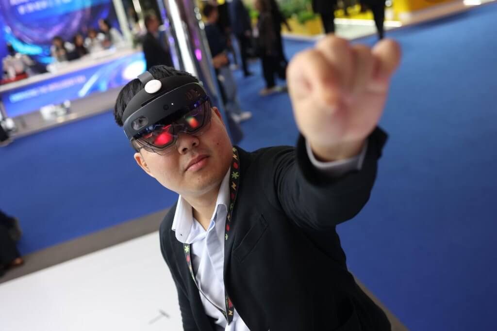 Man wearing AR glasses and headset, reaching forward with his hand at a tech event with blue flooring.