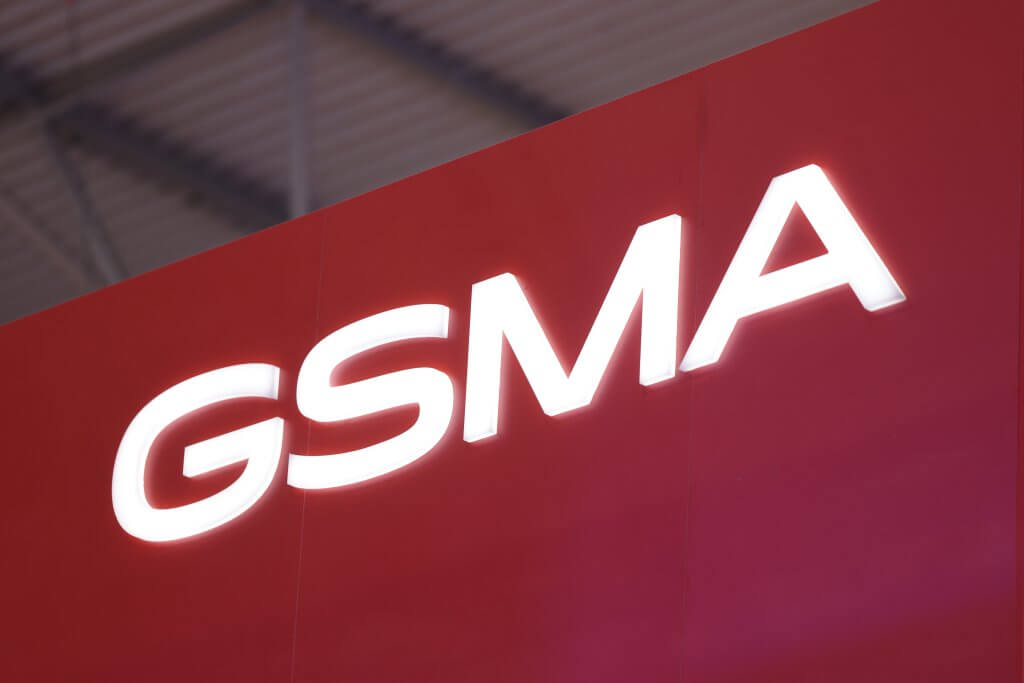 White "GSMA" letters on a bright red background, with a blurred industrial ceiling in the background.