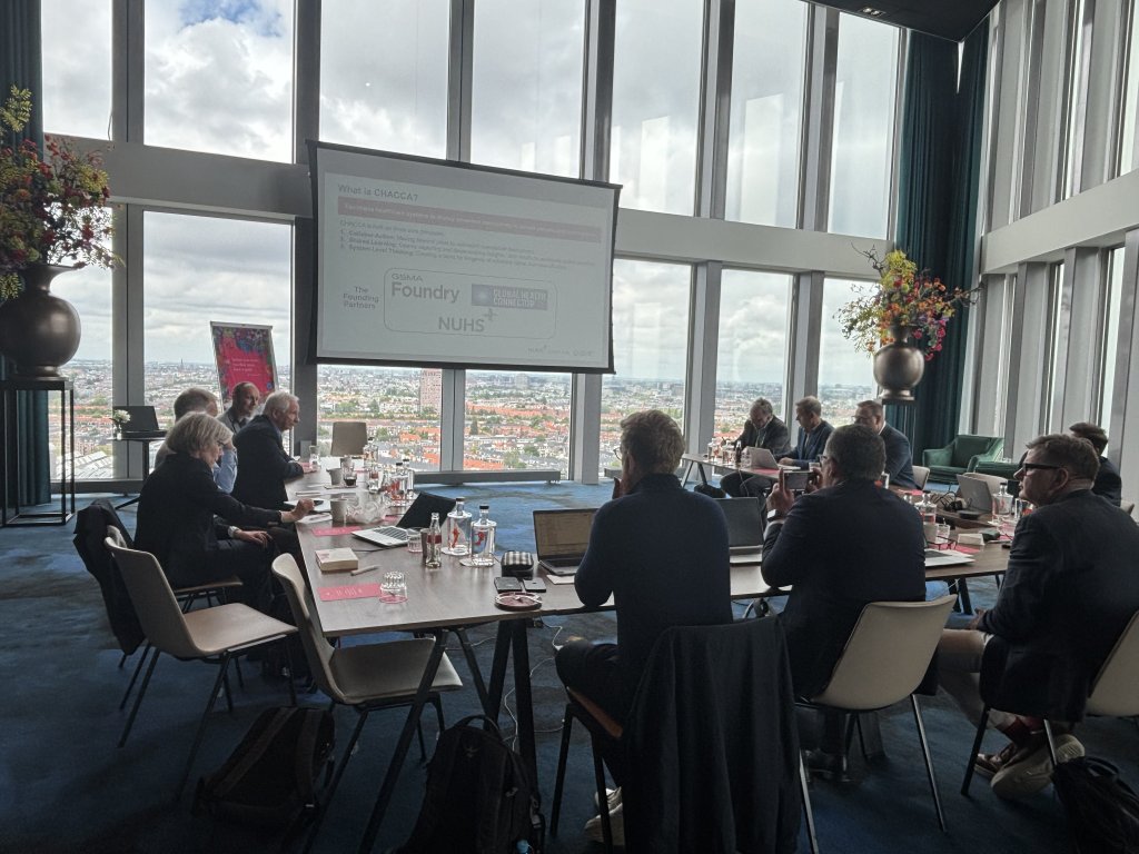 A group of people sit around a conference table in a modern, high-rise room with floor-to-ceiling windows, cityscape visible outside. A presentation is projected on a screen at the front, and large flower arrangements are placed near the windows.