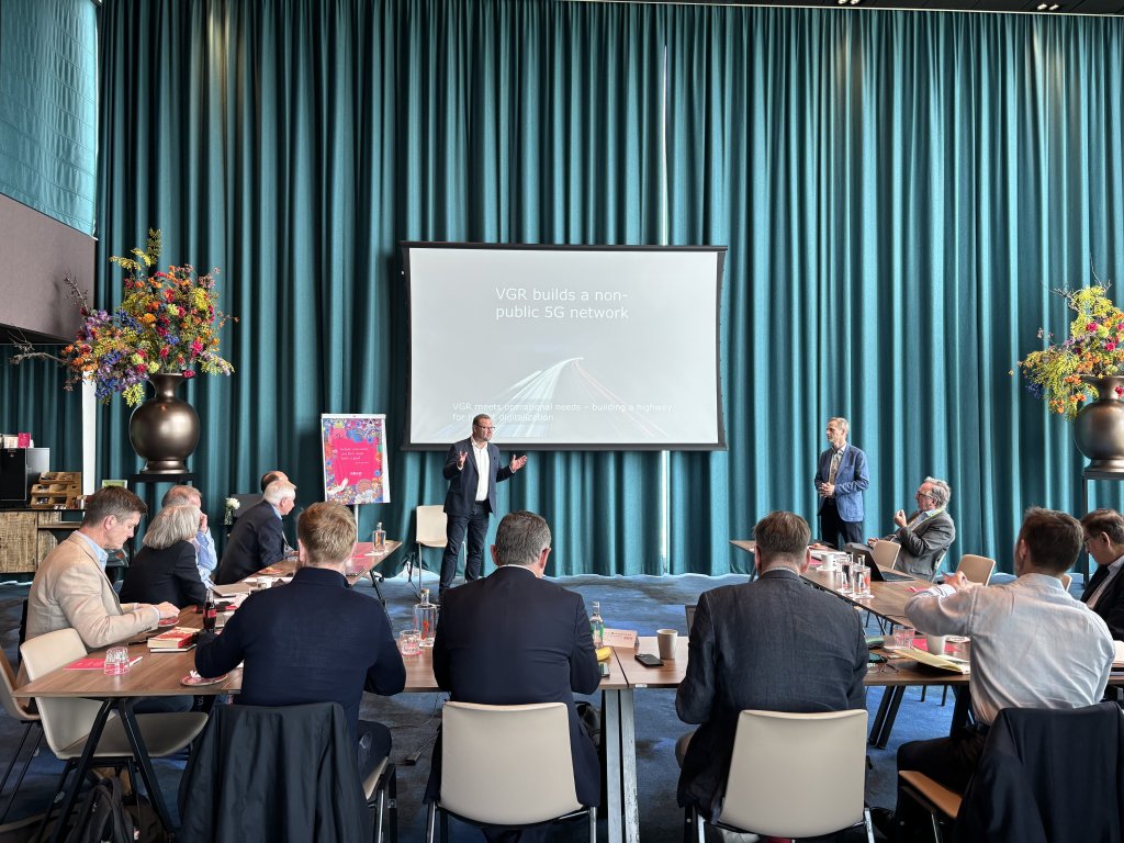 A man stands and presents in front of a projection screen reading “VGR builds a non-public 5G network” to a seated group of people in a conference room. Another man stands nearby. The room has large teal curtains, flower arrangements, and tables with drinks and papers.