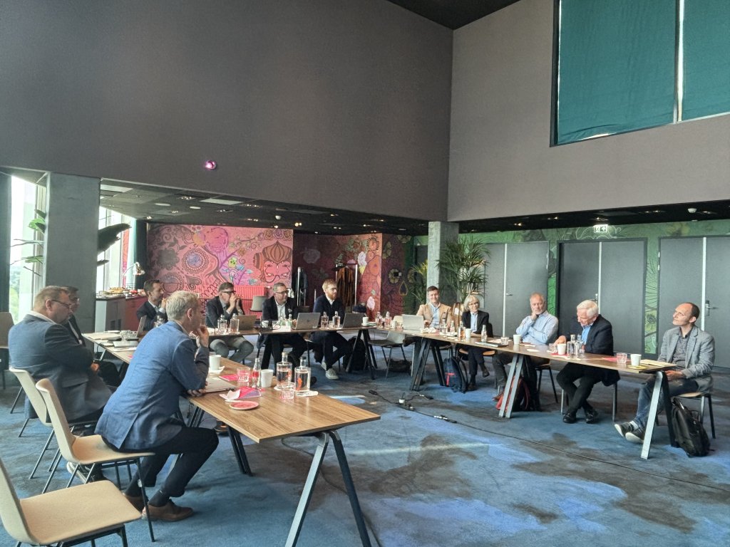 A group of people in business attire sit around a U-shaped table arrangement in a modern meeting room with large windows, bright floral wallpaper, and various drinks and papers on the tables. Some participants are listening while one person speaks.