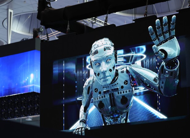 A large digital screen displays a realistic humanoid robot reaching forward with one hand raised, as if touching or pressing against the screen. The background features futuristic blue and white lights, creating a high-tech, sci-fi atmosphere.