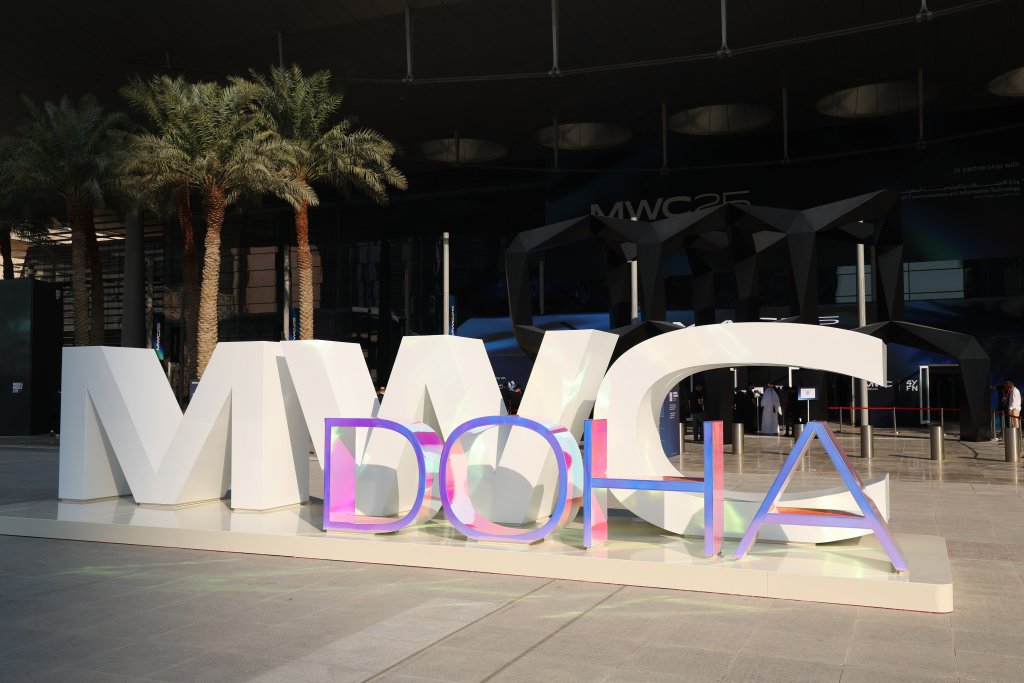 Large white letters spell "MWC" with iridescent "DOHA" letters in front. Palm trees and a modern building entrance are in the background, partially shaded, with "MWC23" displayed above the doorway.