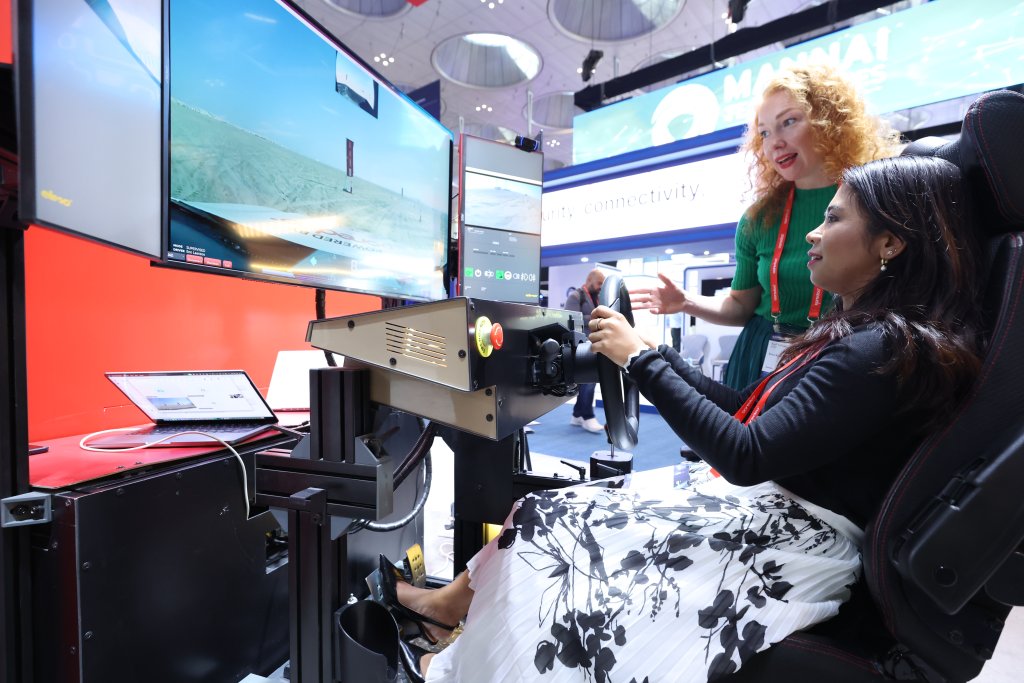 Two women interact at a flight simulator setup. One sits, holding the controls and concentrating on the screen, while the other stands beside her, instructing. Monitors show a flight simulation, and an exhibition environment is visible in the background.