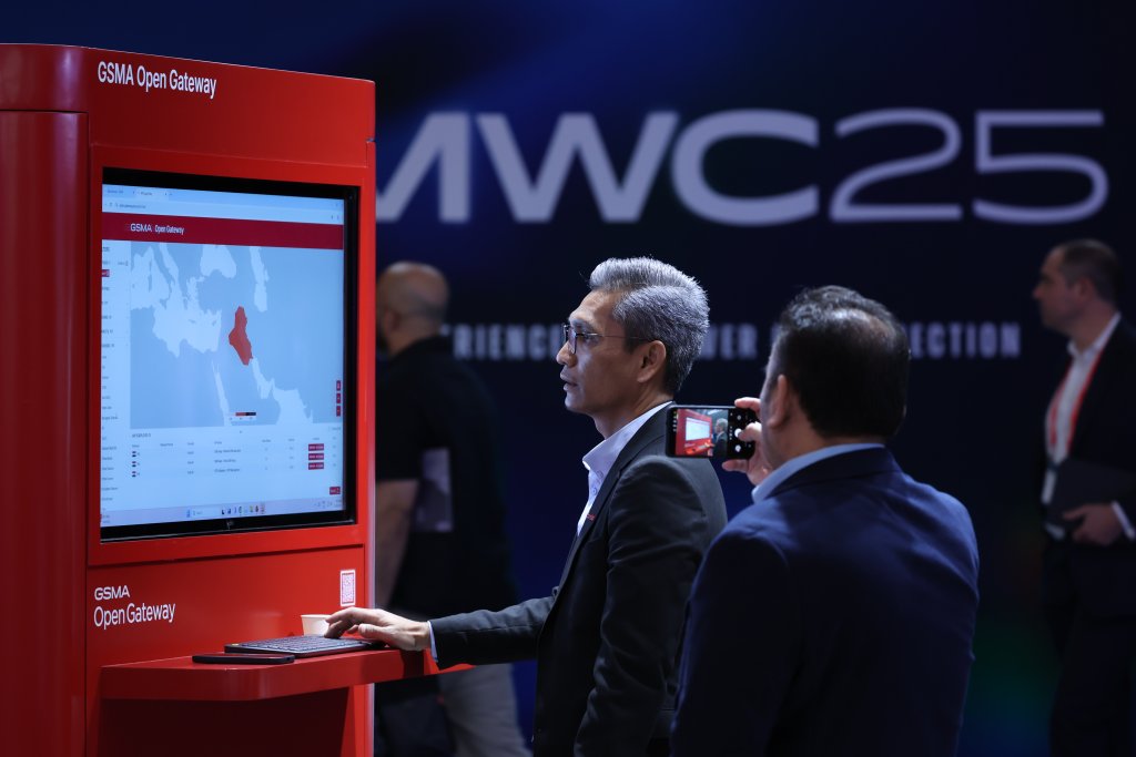 Two men stand at a red GSMA Open Gateway kiosk with a large screen displaying a world map and data. One man uses the touchscreen, while the other films or photographs the scene. In the background, “MWC25” is visible on a dark blue wall.