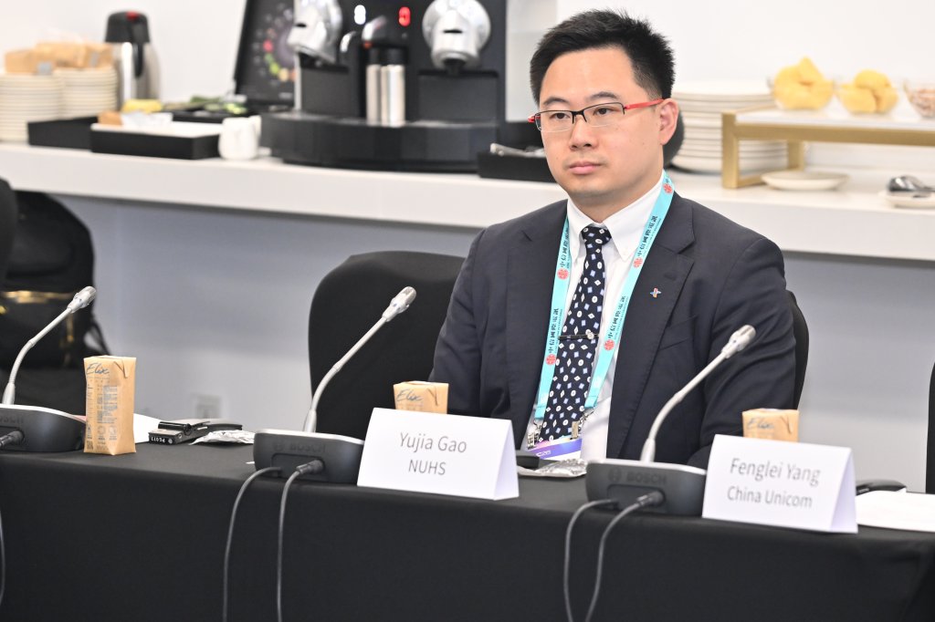 A man in a suit and glasses sits at a conference table with microphones, drinks, and name cards that read “Yujia Gao NUHS” and “Fenglei Yang China Unicom.” Behind him are coffee machines, plates, and pastries on a counter.