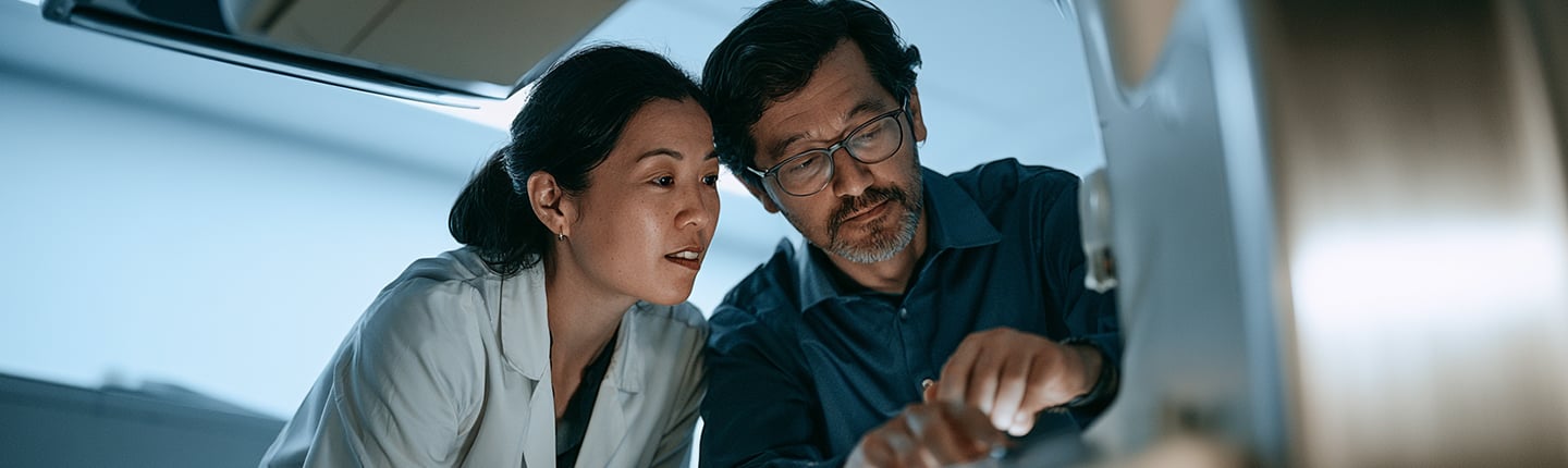 Two people, a woman in a white lab coat and a man with glasses and a beard, closely examine equipment in a lab or medical setting, illuminated by soft overhead lighting. They appear focused and engaged in a technical or scientific task, exemplifying the spirit of collaboration found within a Connected Healthcare Content Hub.