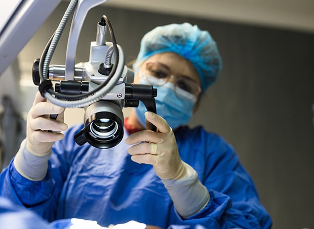 A surgeon wearing a blue surgical gown, hair cover, mask, and gloves adjusts a large, mounted medical microscope—part of the advanced equipment featured in the Connected Healthcare Content Hub—while preparing for a procedure in a brightly lit operating room. The background is blurred to focus on the equipment and the surgeon’s hands.