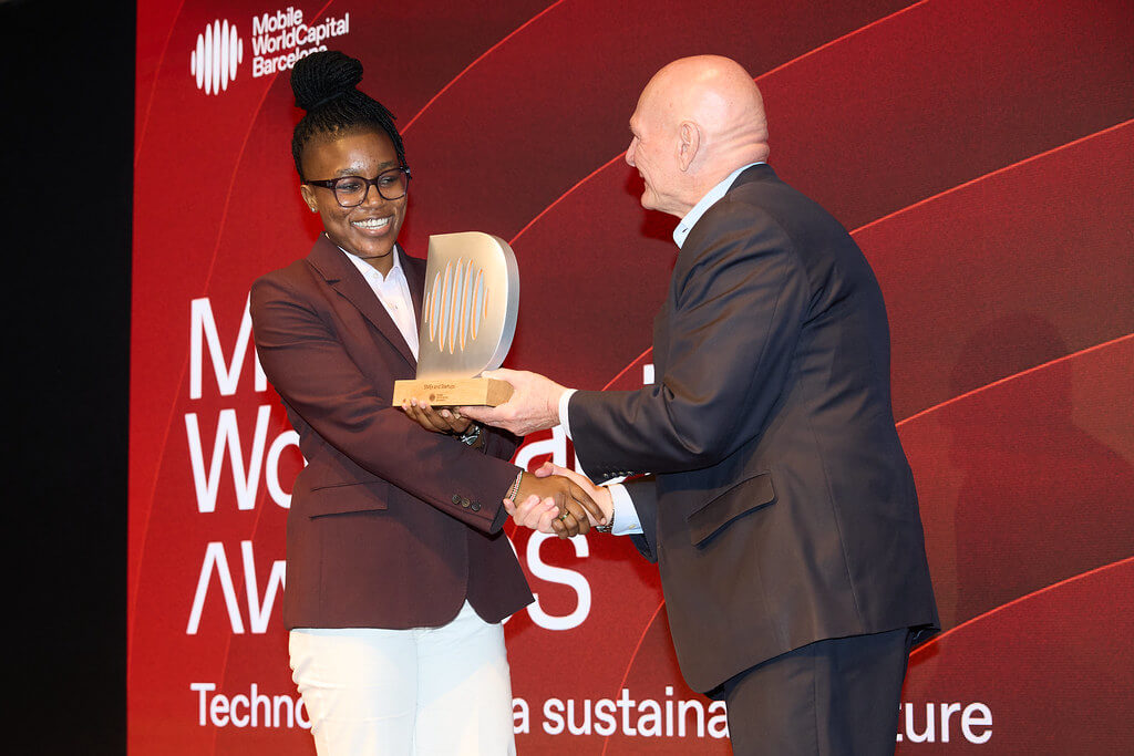 A woman in glasses and a maroon blazer smiles while receiving an award from a bald man in a suit on stage at the Mobile World Capital Awards in Barcelona. The red background displays event branding and white text.