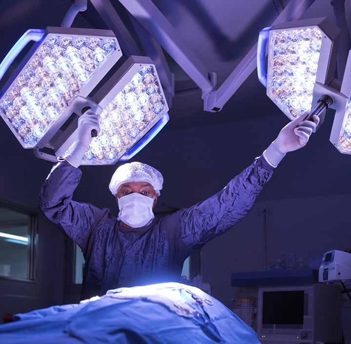 A surgeon in blue scrubs, mask, and hair cover adjusts two large surgical lights above an operating table, preparing for surgery in a dimly lit operating room equipped with advanced medical technology that supports Connected Healthcare solutions.