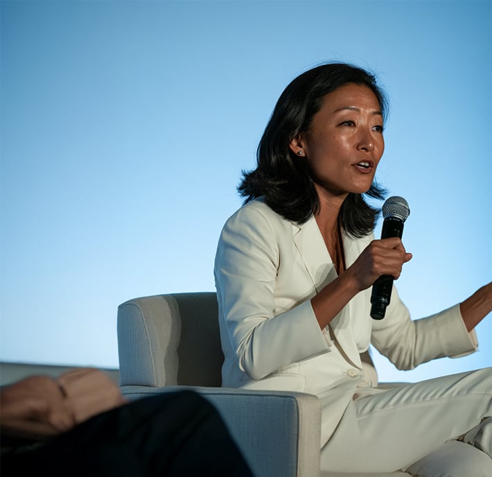A woman with medium-length dark hair, wearing a white suit, sits in a chair speaking into a handheld microphone with one hand raised. She is engaged in conversation about Connected Healthcare against a soft blue background, with another person’s hand and arm visible in the foreground.