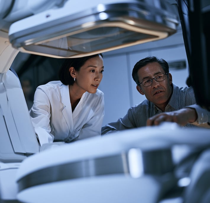 Two professionals examine advanced medical equipment, demonstrating the impact of Connected Healthcare in a modern clinical or laboratory setting. The woman wears a white lab coat and the man wears glasses and a striped shirt. Both appear focused as they collaborate amid metallic and white surfaces, highlighting how Connected Health technologies enhance their work environment.